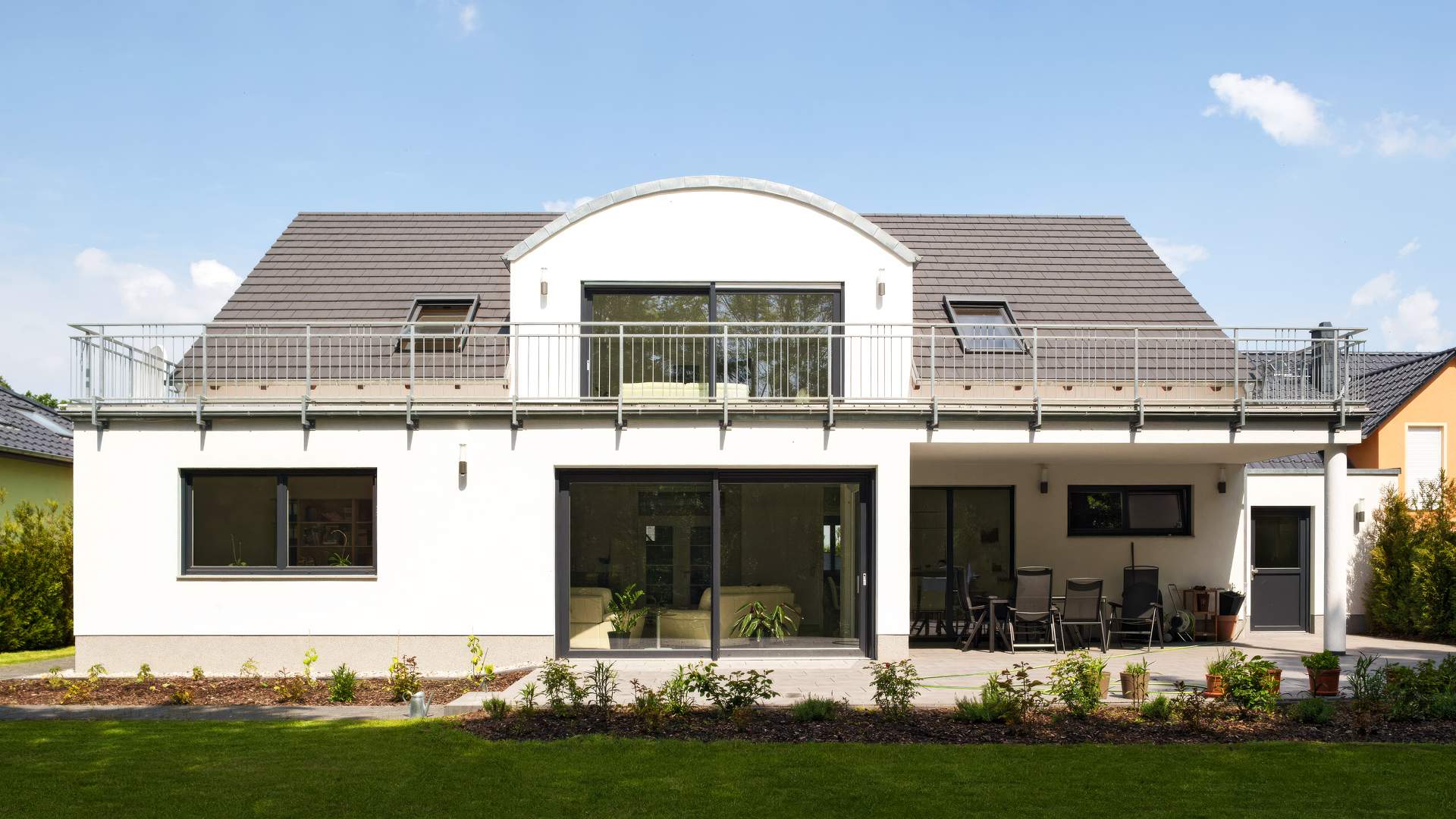 Moderne Hausrückseite mit großer Fensterfront, weitläufigem Balkon mit Metallgeländer und gepflegtem Garten unter blauem Himmel